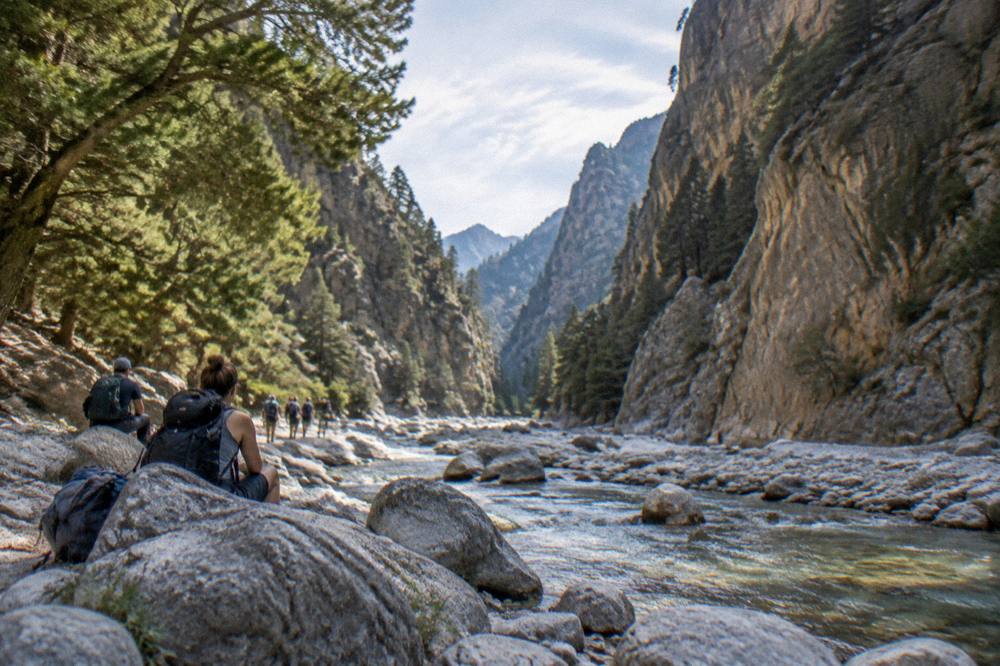 Hikers with backpacks resting beside a stream in a wide stony gorge