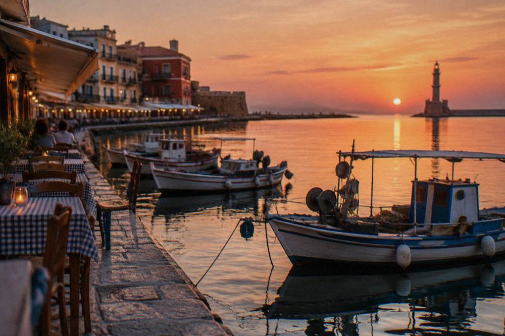A seafront promenade at dusk with taverna tables and fishing boats moored in a small marina
