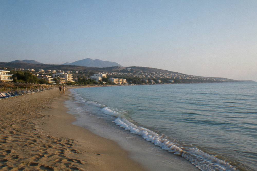 A long sandy beach backed by low-rise hotels and green hills under a clear sky