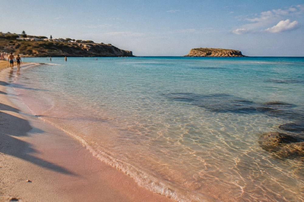 A wide shallow lagoon with pale pink sand and turquoise water between a headland and an island