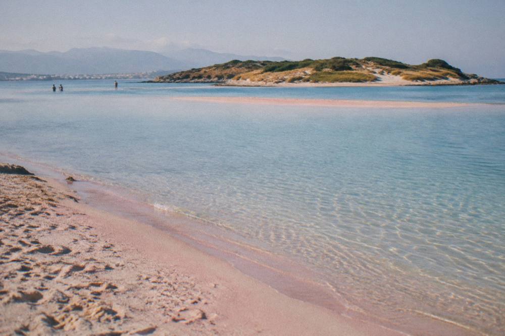 A shallow lagoon between a pale pink sandbar and a small island with dune grasses