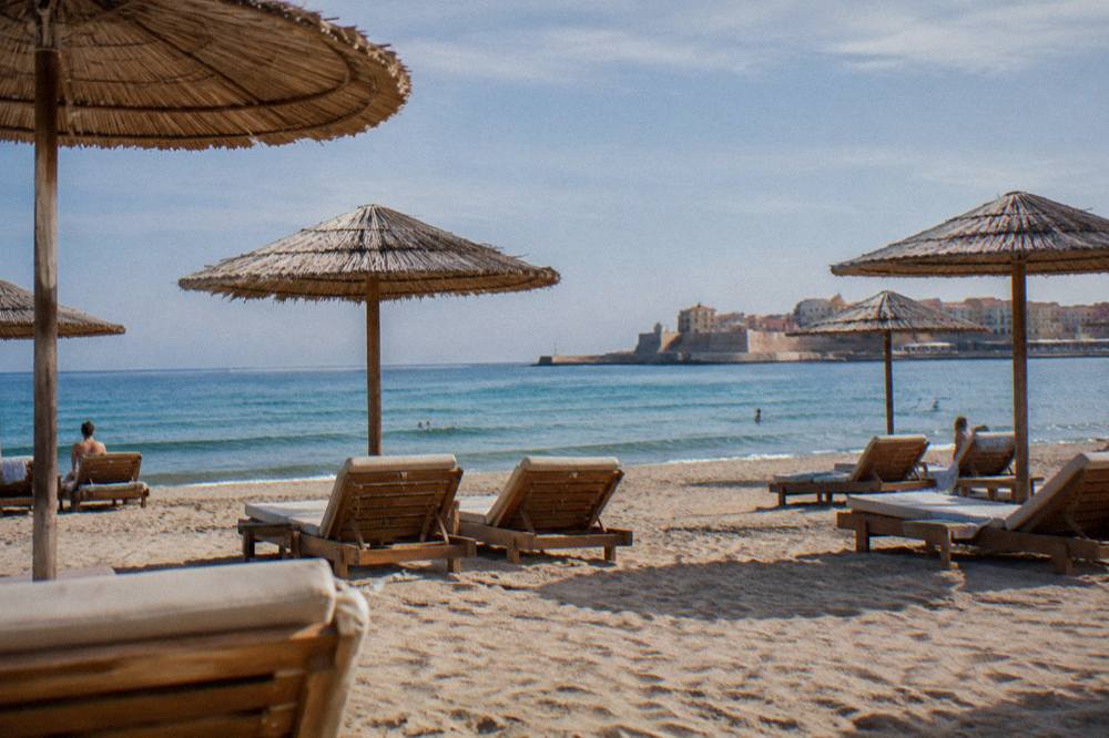 Sun loungers on sand under umbrellas with a calm blue sea and clear sky behind them