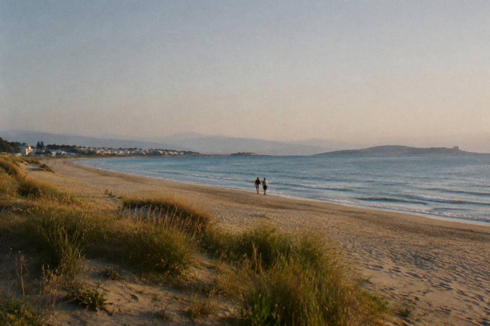 A long open sandy beach with low dunes and grasses facing a calm sea at late afternoon