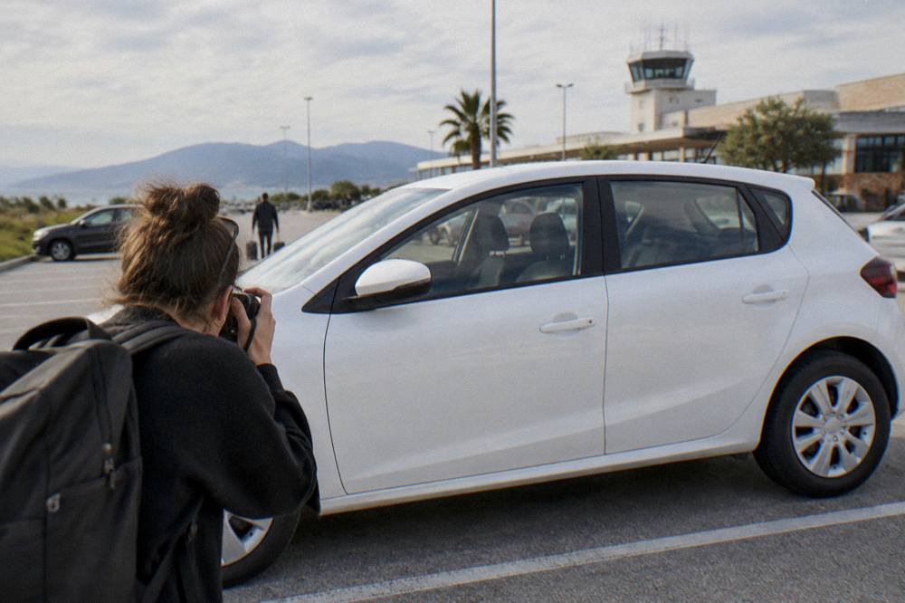 A person photographing the side panel of a white hire car in a car park