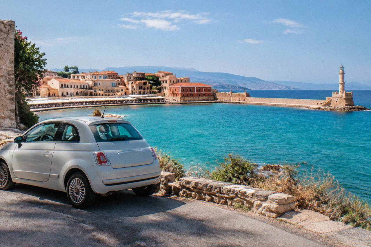 A small rental car parked on a coastal road above a turquoise bay in western Crete