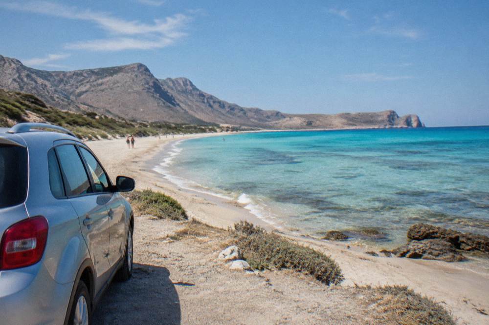 A parked car beside a long sandy beach with shallow turquoise water at Falassarna