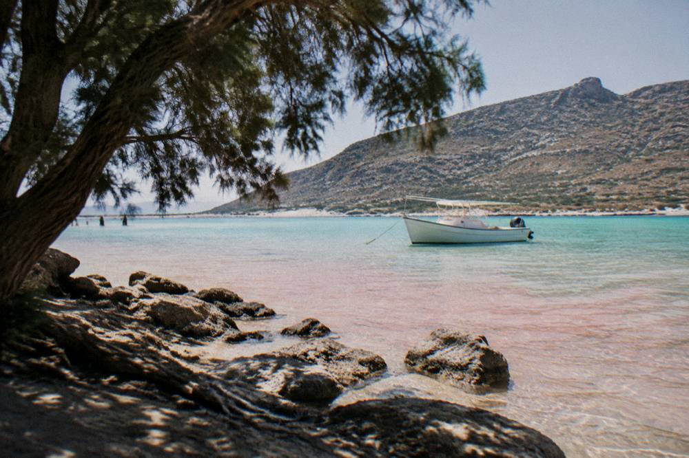 A white boat moored in shallow pink-tinged water at Balos lagoon with hills behind