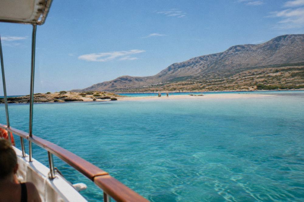 A shallow turquoise lagoon with a sandy spit and hills behind, seen from a boat