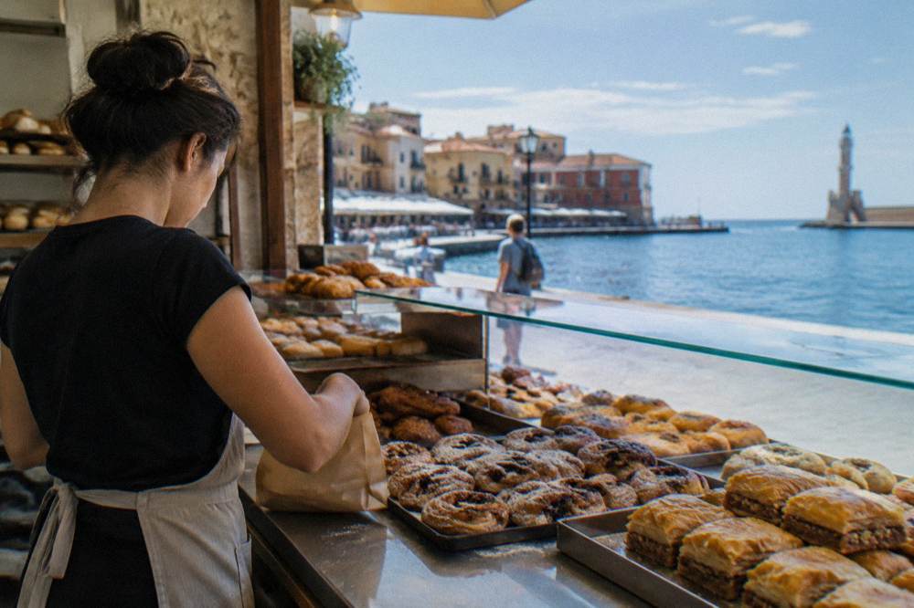A bakery counter in Chania with trays of pastries and a server wrapping an order
