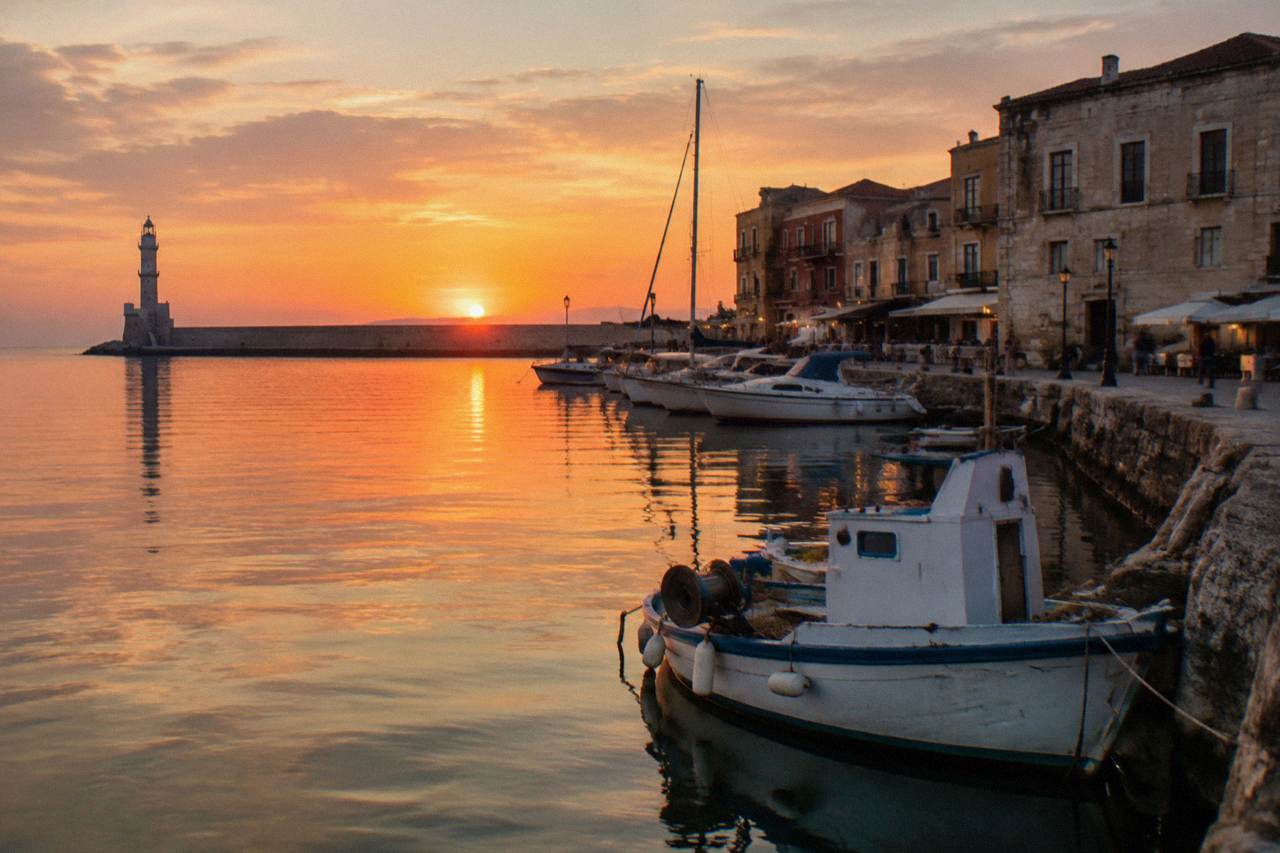 Evening light over a Cretan harbour front with moored boats and stone buildings along the quay