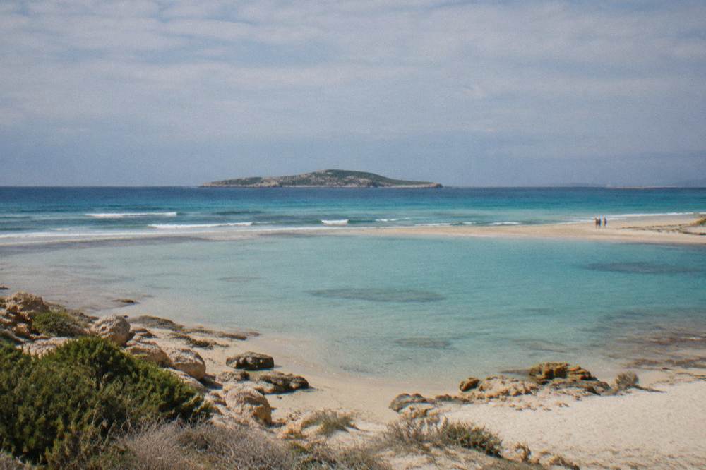 A shallow turquoise lagoon behind a sandbar with a small island on the horizon