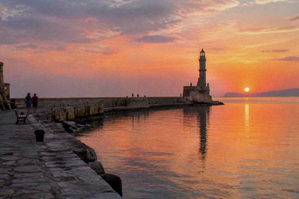 A stone harbour mole curving out to a lighthouse under a pink and blue evening sky