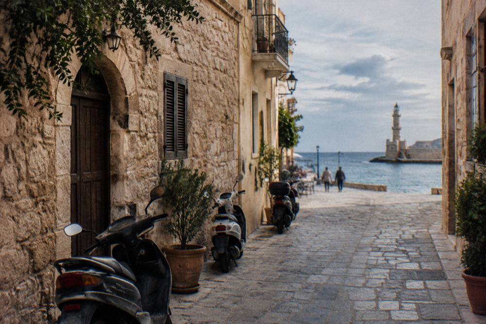 Narrow old town street with stone buildings and parked scooters near a harbour