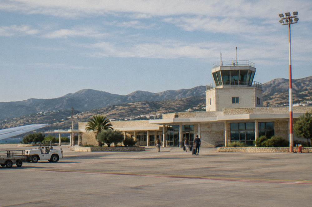 A small airport terminal building under a bright Mediterranean sky with hills behind