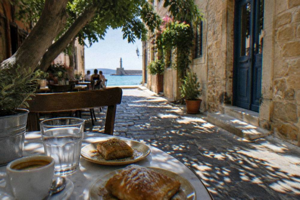 A cafe table with coffee and pastries in a shaded Chania lane