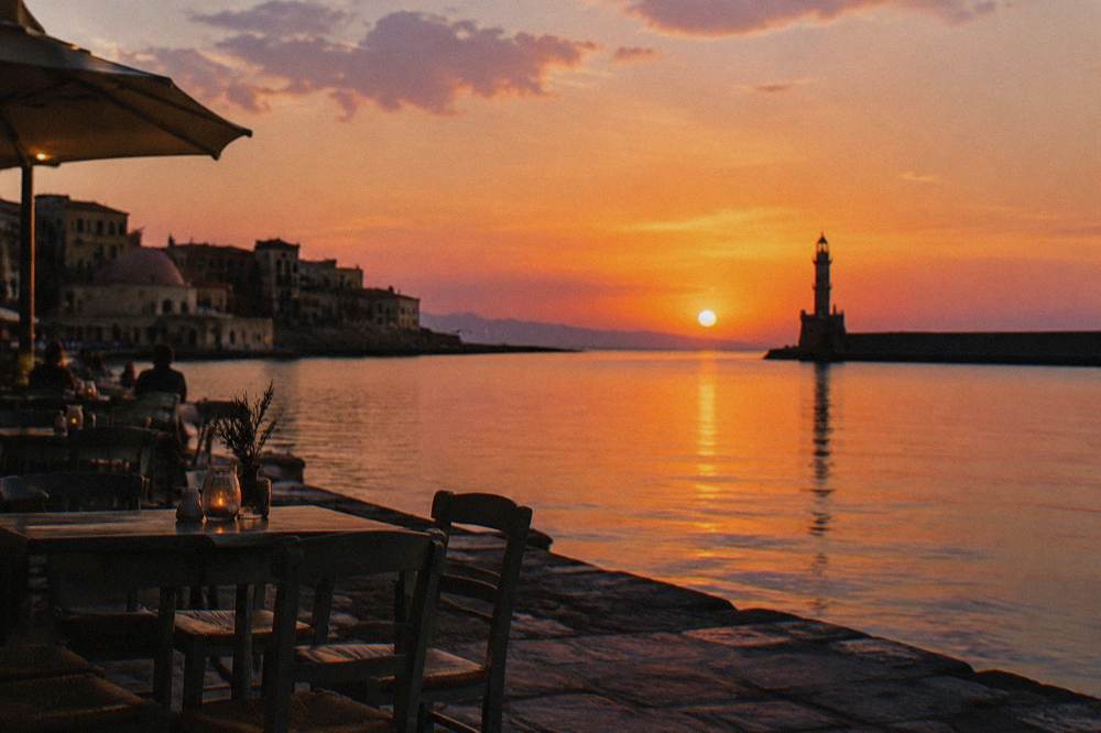 Sunset over Chania harbour front with cafe tables and the silhouette of the lighthouse