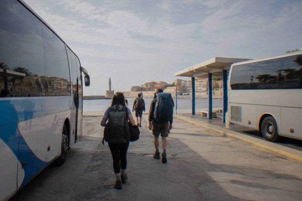 Travellers with backpacks walking beside buses at a small Greek bus station