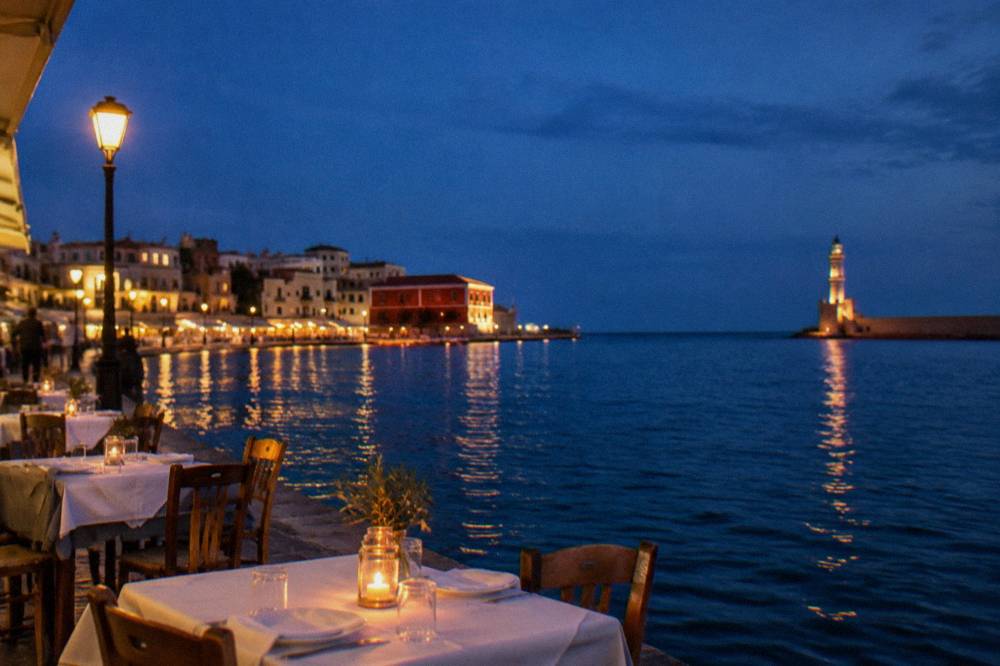 Outdoor restaurant tables set along the lit harbour quay at dusk in Chania