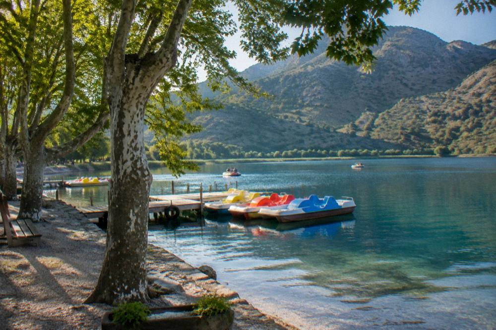 A calm freshwater lake with pedalos moored at a jetty and green hills at Lake Kournas