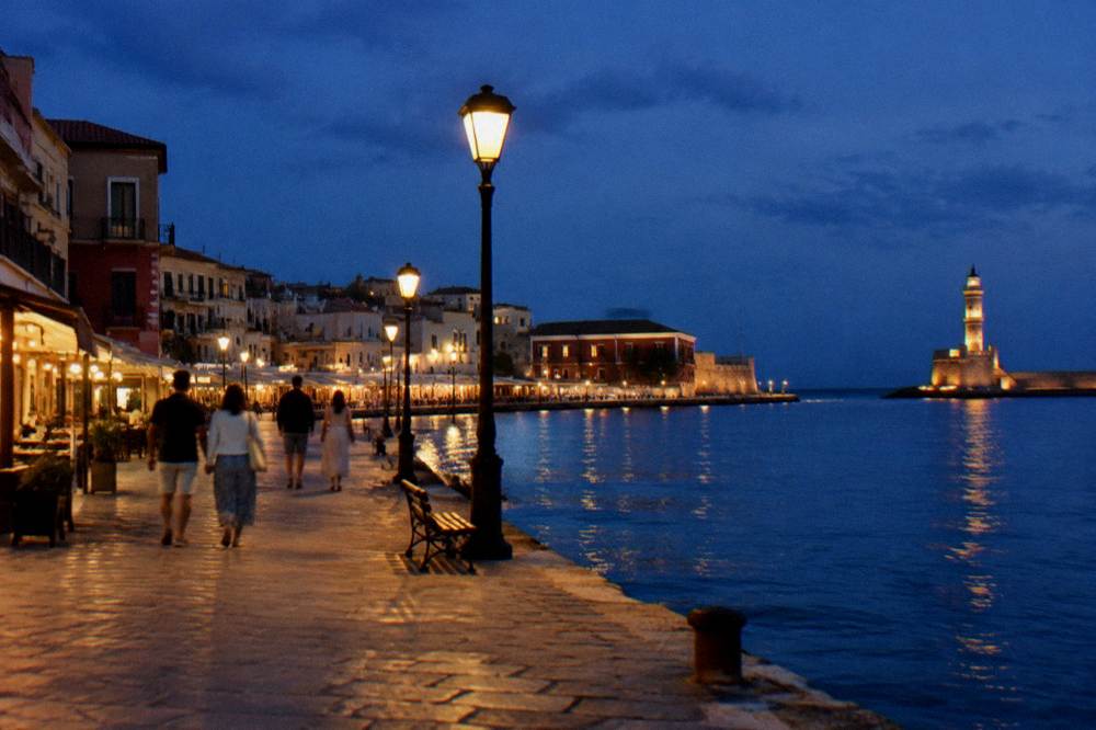 A small group walking along the lit harbour quay in Chania at blue hour