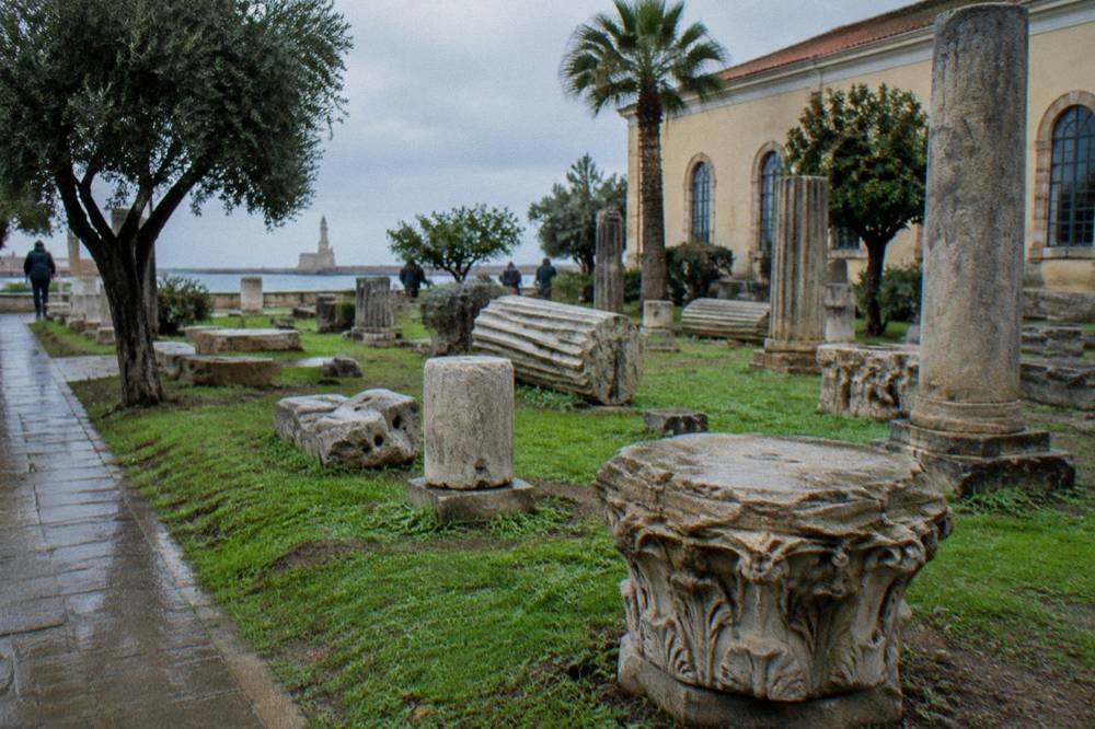 Weathered ancient stone fragments laid on grass in a museum garden in Chania under a grey sky