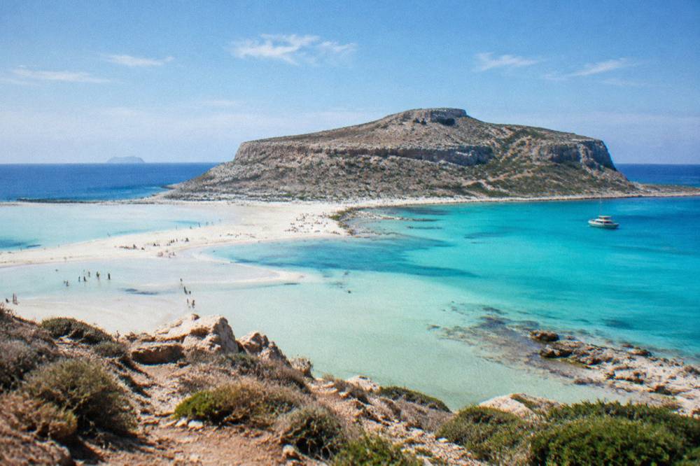 A shallow turquoise lagoon of pale sand beneath a rocky headland at Balos