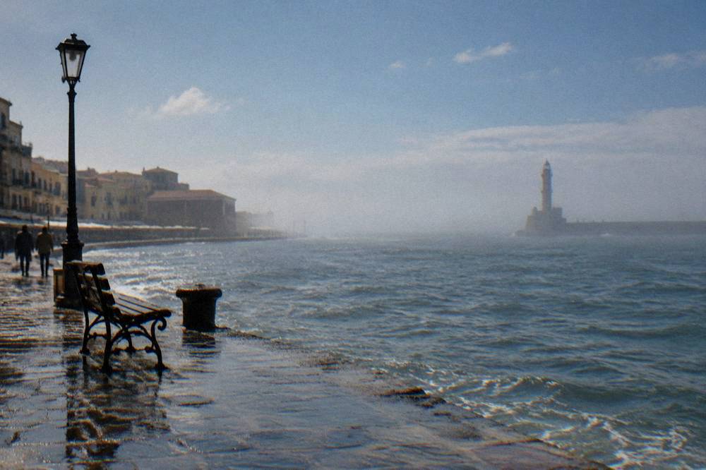 Wet harbour front in Chania with the lighthouse faint in mist across grey water